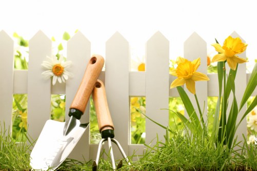 A close-up view of a well-maintained garden in Leyton, featuring a white picket fence in the background, with yellow daffodils and white daisies blooming along the fence line. In the foreground, there is a patch of lush, green grass with a slightly textured surface. Gardening tools, including a trowel and a hedge trimmer with wooden handles, rest on the grass, indicating ongoing or recent garden maintenance. The garden area appears tidy and healthy, with natural sunlight illuminating the scene, highlighting the vibrant colors of the flowers and the fresh greenery. This outdoor space showcases typical features of a landscaped garden in Leyton that could benefit from professional gardening services focused on lawn care, planting, and garden tidying, aligned with local gardening needs and aesthetics.