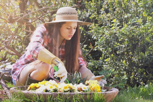 A young woman wearing a wide-brimmed straw hat, a red plaid shirt, and gardening gloves is carefully planting or tending to colorful flowers within a raised garden bed. The garden bed is embedded in a lush, well-maintained residential backyard featuring a neatly mowed green lawn and dense, leafy hedges and shrubs in the background. The scene is illuminated by natural daylight, suggesting a bright, clear day, with sunlight filtering through the foliage, creating a warm and inviting outdoor environment. The garden area appears organized and healthy, reflecting professional gardening practices typical of Leyton garden maintenance services, with visible soil, vibrant flower blooms, and a natural, landscaped setting suitable for outdoor leisure and horticultural care.